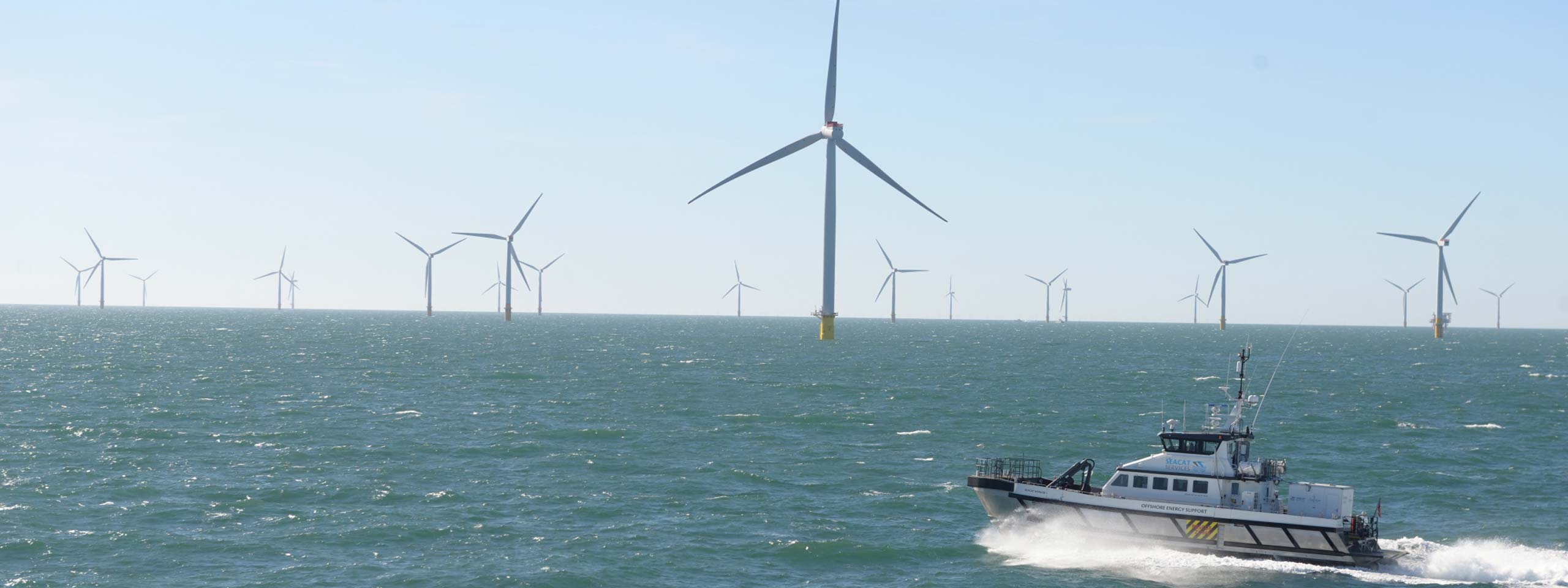 A boat navigating through waves near offshore wind turbines in a clear sky.
