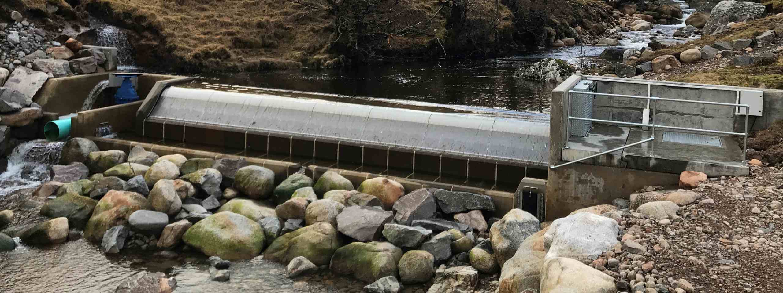 A hydroelectric dam beside a river, with large rocks and a blue turbine visible in the water.