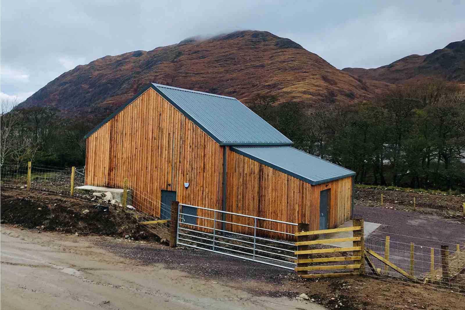 A wooden building with a metal roof, set against a mountain backdrop. The landscape is rocky and partially cloudy.