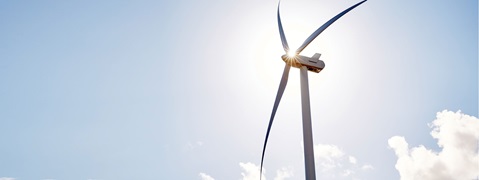 A tall wind turbine with three blades, set against a clear blue sky and bright sunlight.