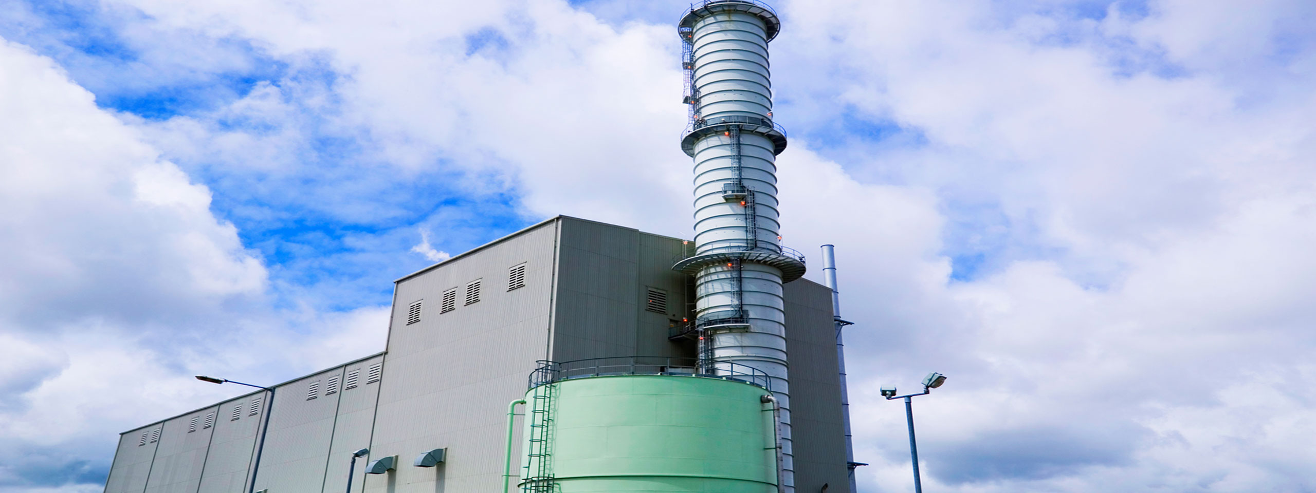 A power plant structure with a tall metal stack, surrounded by clouds and blue sky.