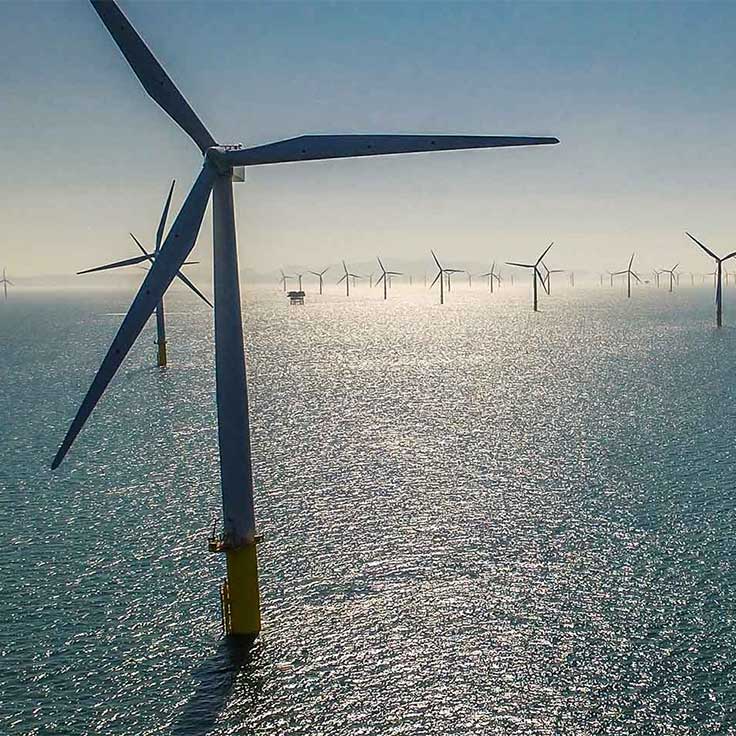 Aerial view of offshore wind turbines in calm waters under a clear sky, reflecting sunlight on the sea surface.
