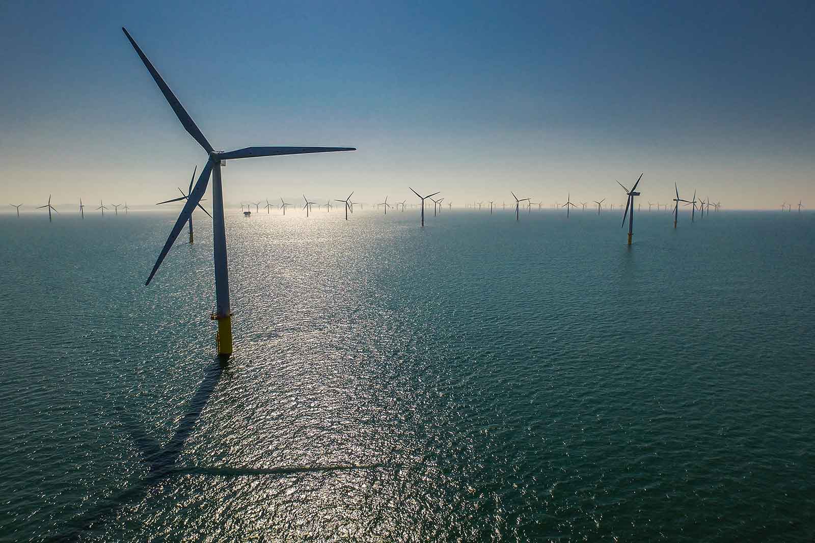 Aerial view of wind turbines in the ocean, with reflections on the water and a clear sky in the background.