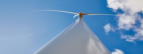 A tall wind turbine viewed from below against a bright blue sky with scattered clouds.
