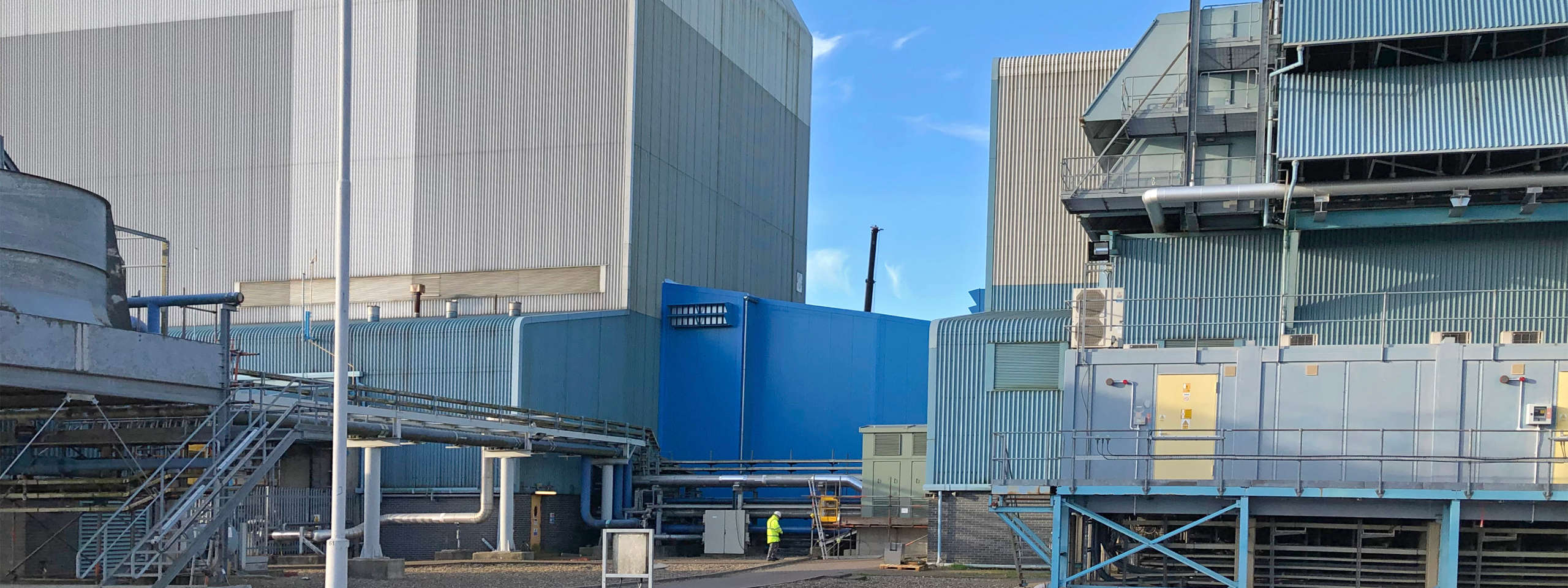 Industrial facility with large metal structures, pipes, and a person in a high-visibility jacket.