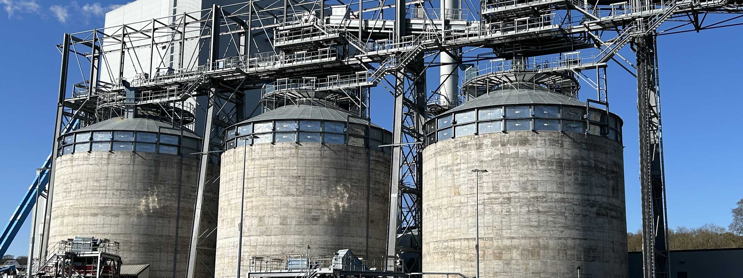 A large industrial facility featuring three cylindrical silos, surrounded by metal structures and a clear blue sky.