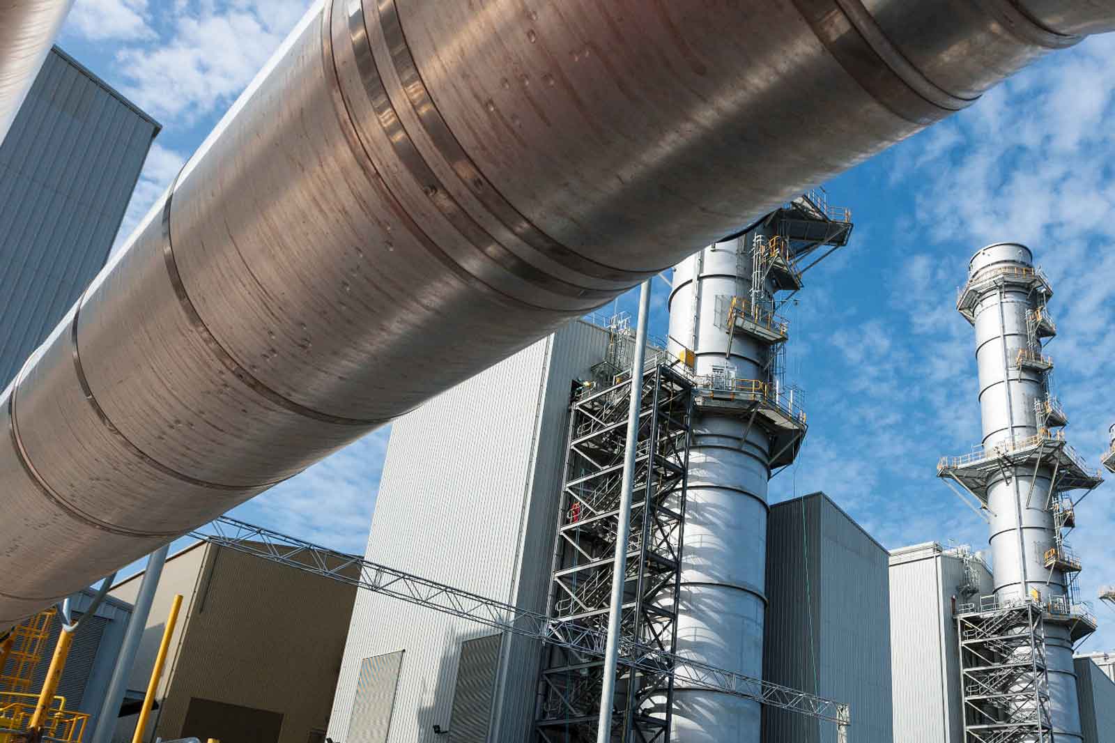 A close-up of large steel pipes at an industrial site with smoke stacks visible under a blue sky.