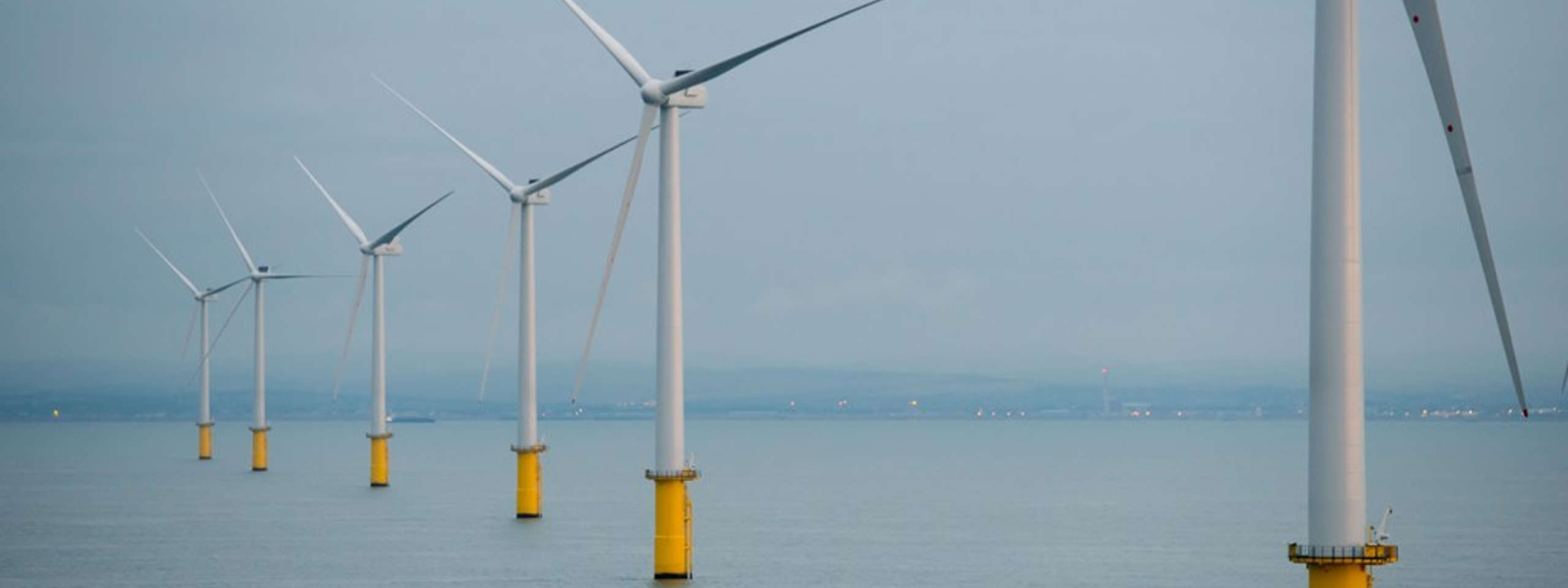 Offshore wind turbines standing tall in the ocean, with yellow foundation visible against a cloudy sky.