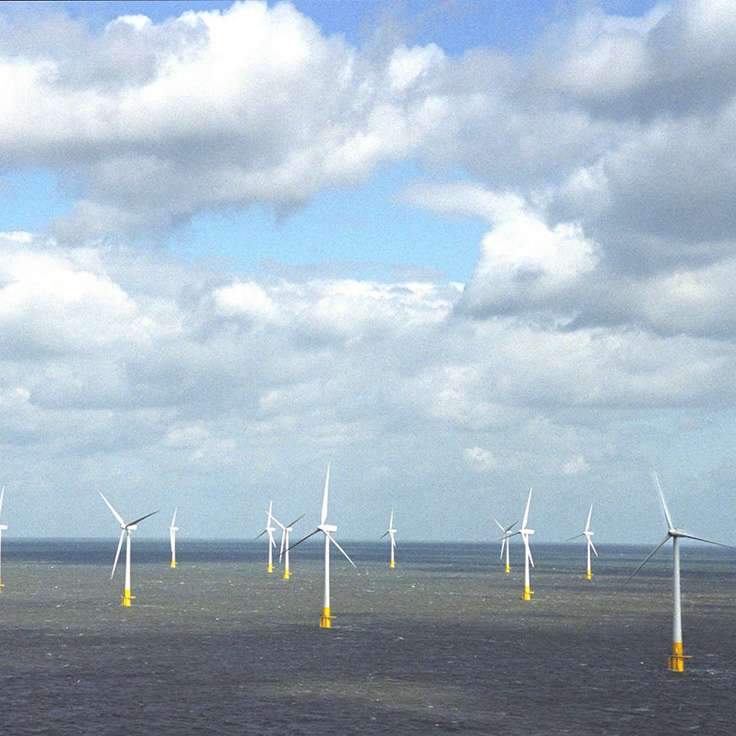 Offshore wind turbines stand tall in the ocean under a partly cloudy sky. The sea reflects the sky's hues.