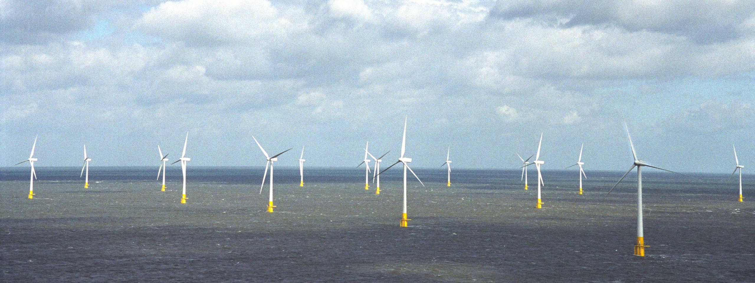 A wide view of offshore wind turbines standing in the ocean under a cloudy sky.