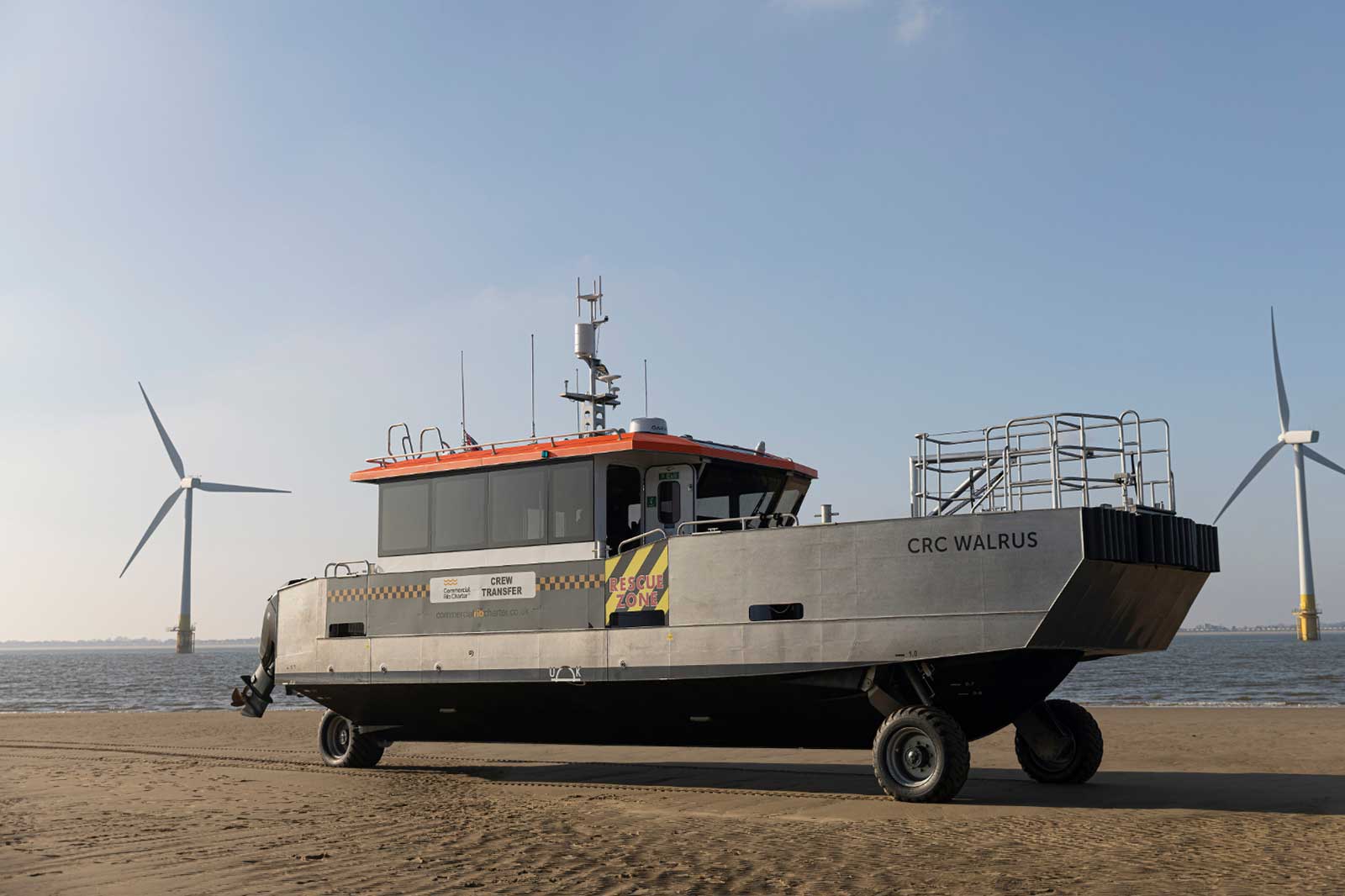 A crew transfer vessel named CRC Walrus stands on sandy shore near wind turbines, showcasing a modern design and wheels.