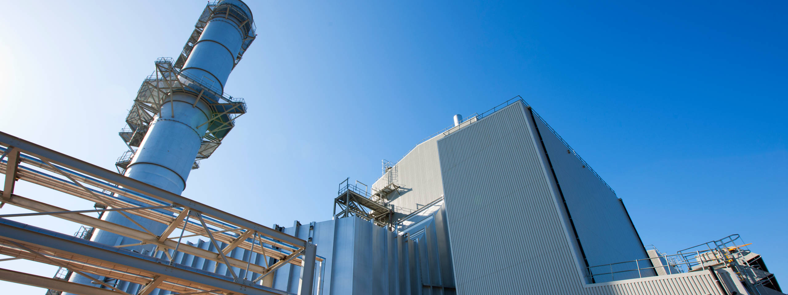 A power plant featuring a tall chimney and metallic structures against a clear blue sky.