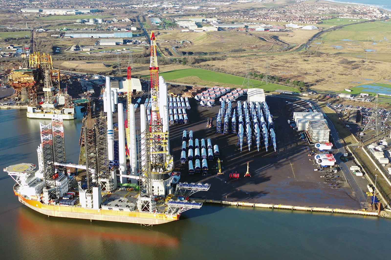 Aerial view of a harbour with ships, wind turbine parts, and industrial structures. Green fields and cityscape in the background.