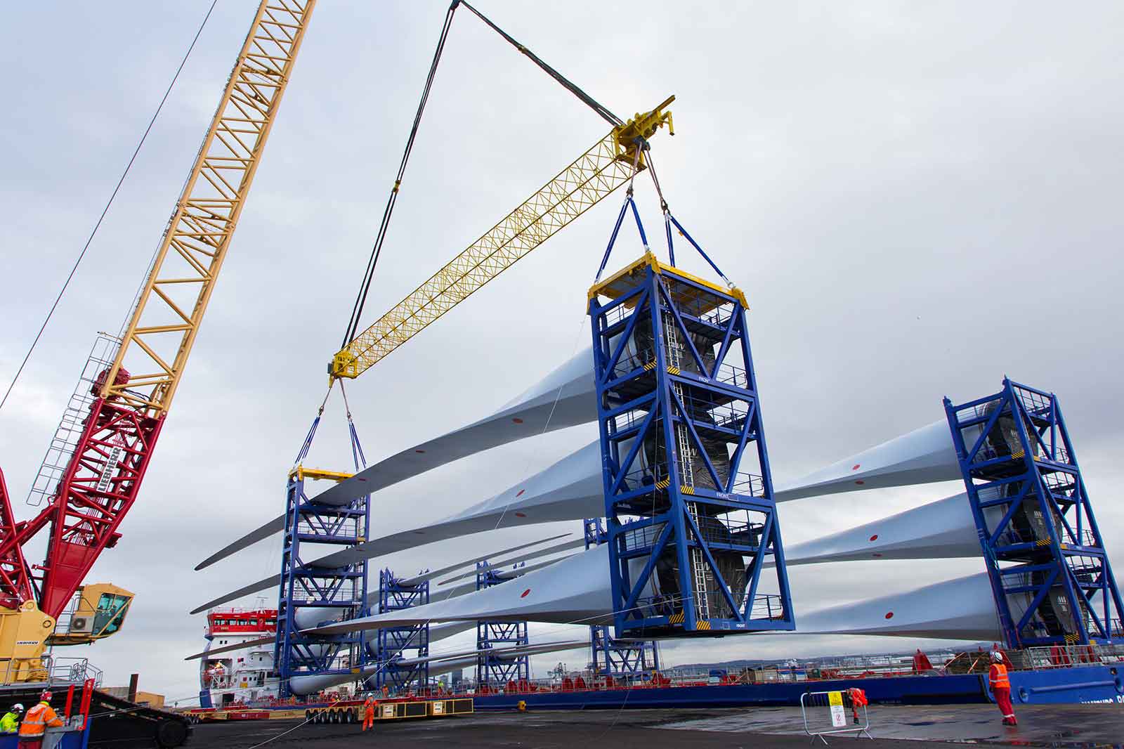 A crane lifts large wind turbine blades at a port. The scene features cranes, workers, and cloudy skies.