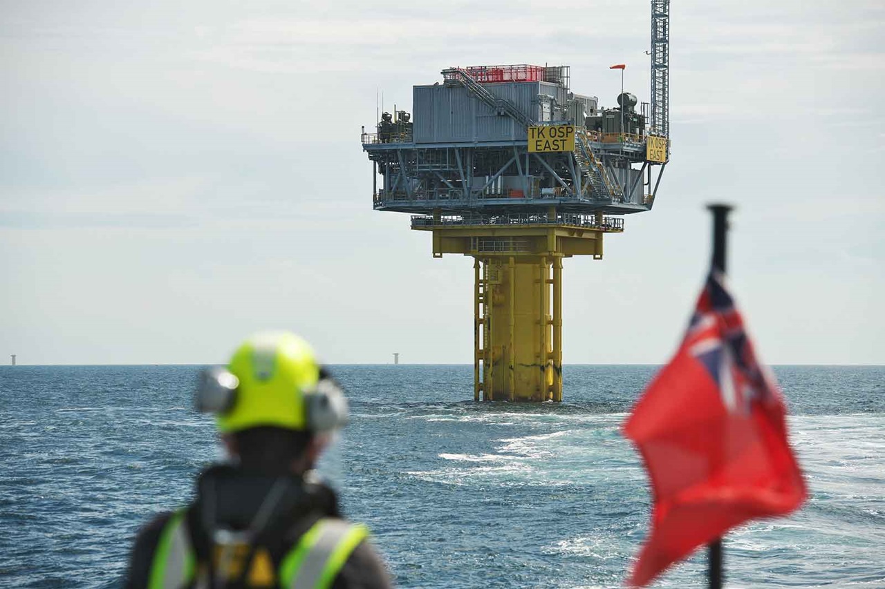 A worker in safety gear observes an offshore oil platform in the sea, with a red flag flying nearby.