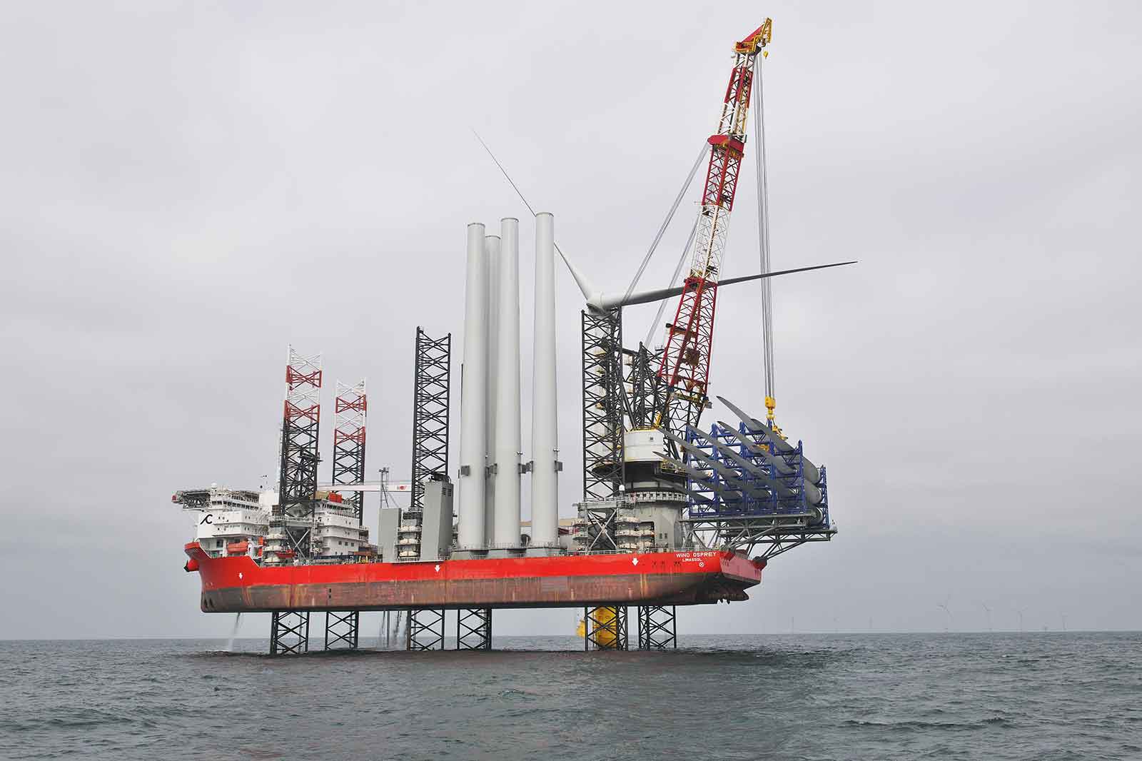 A red and grey offshore vessel with a large crane is installing wind turbine components in choppy waters under a cloudy sky.