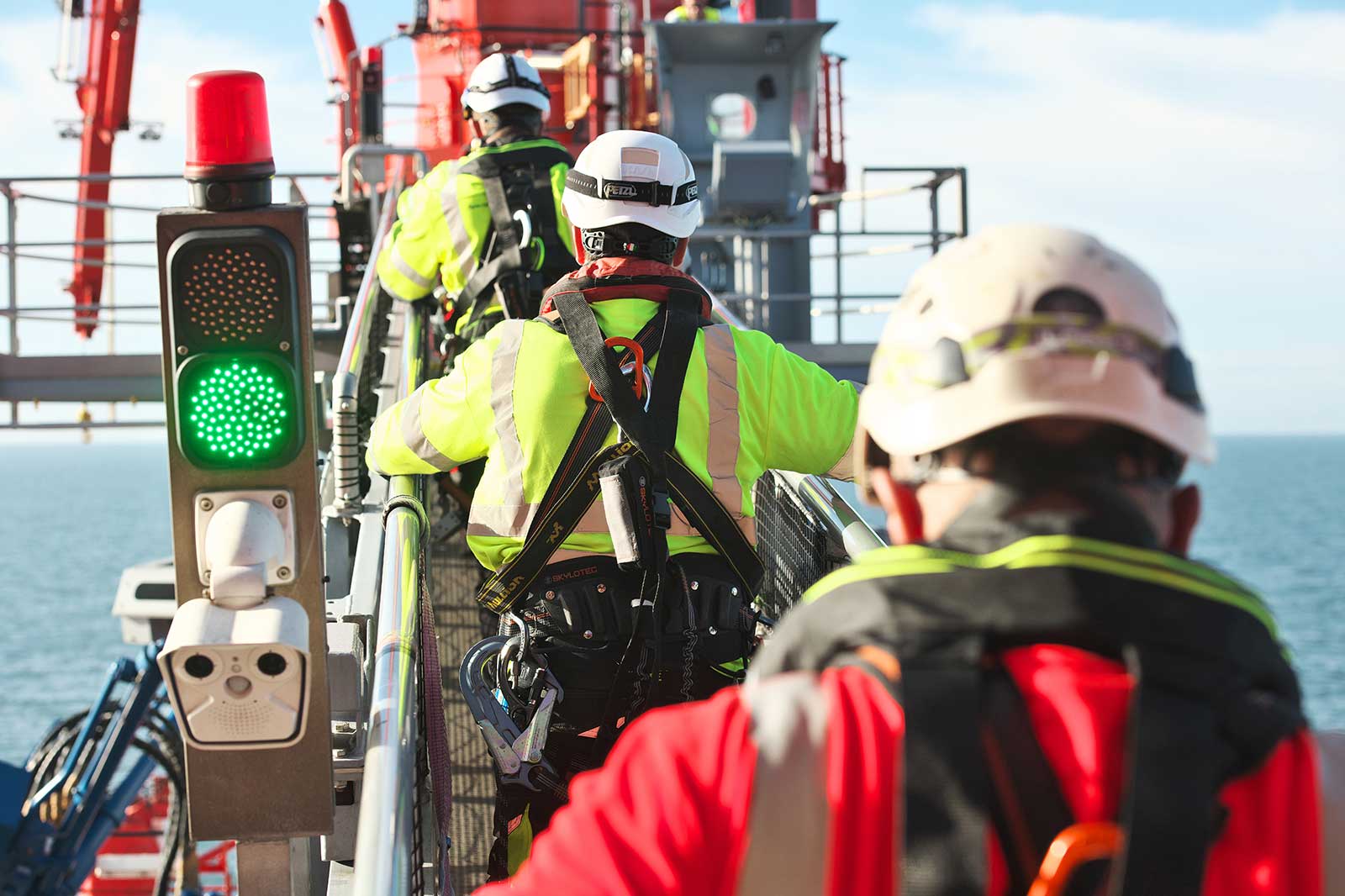 Workers in safety gear ascend a metal walkway near green traffic signal above the sea, with cranes in the background.