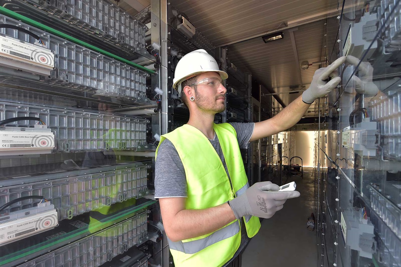 A technician in a safety vest and gloves operates equipment in a storage facility filled with stacked batteries.