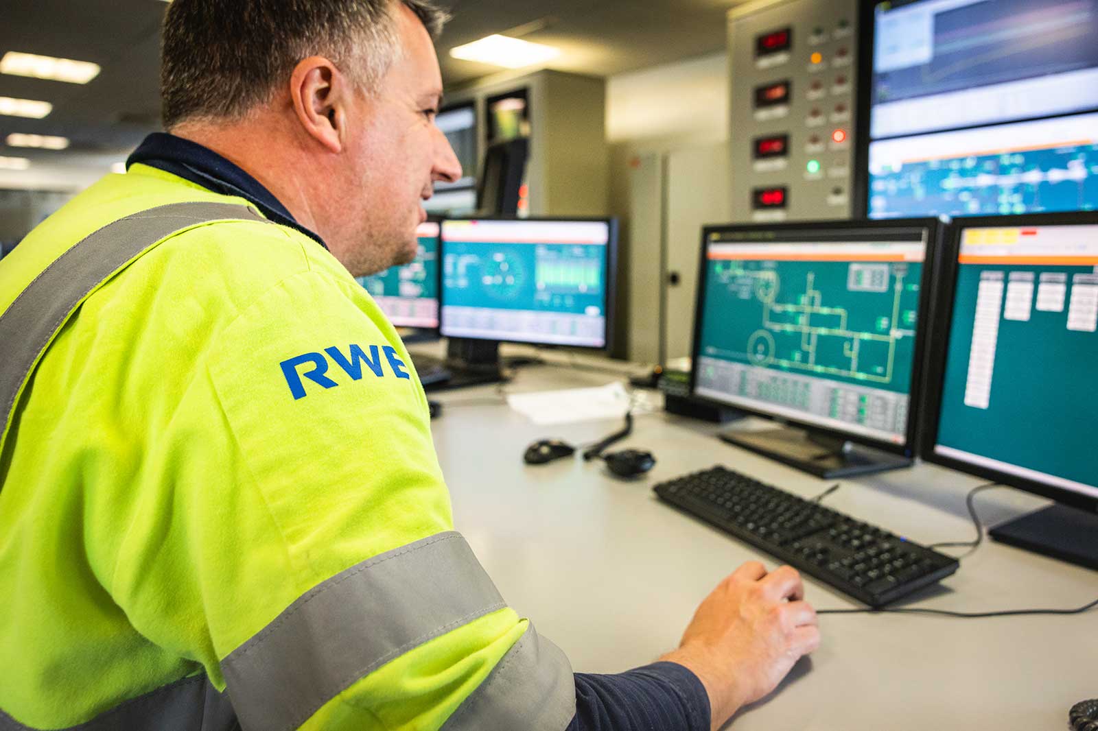 A person in a high-visibility jacket operates multiple computer screens in a control room setting.