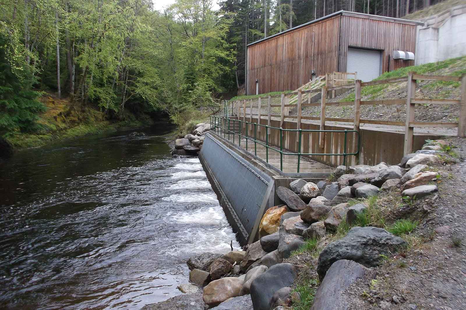 A river flows beside a wooden structure with a concrete water barrier and surrounding rocks, framed by lush greenery.