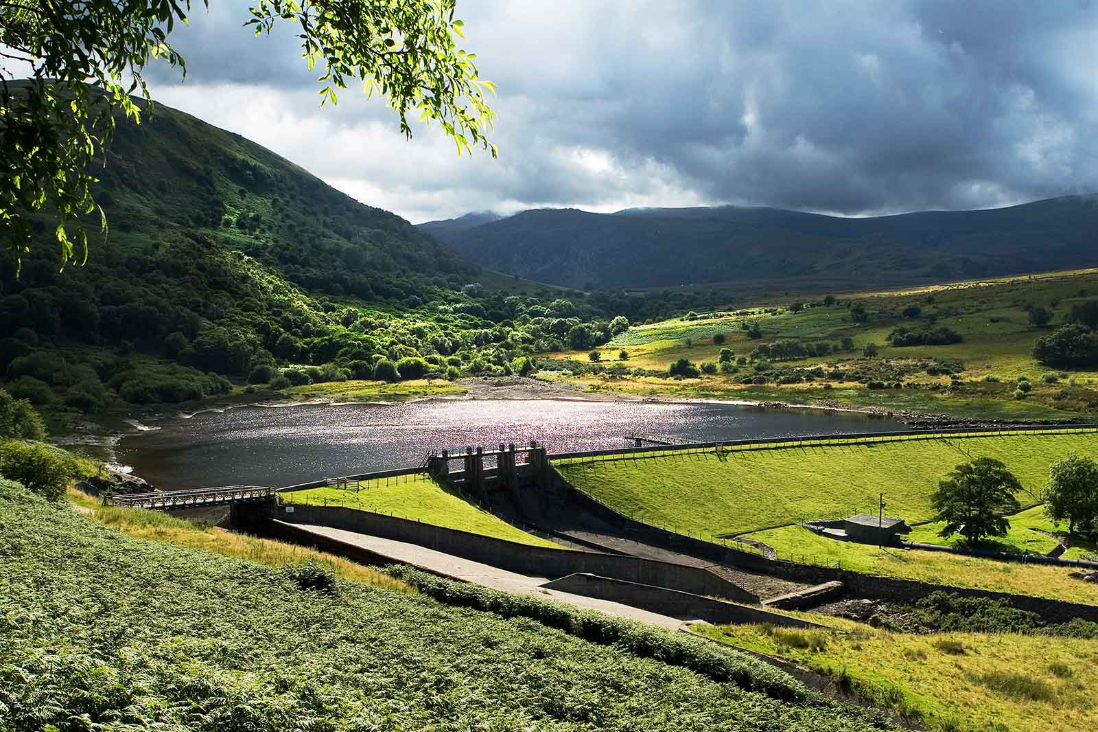 A scenic view of a reservoir surrounded by lush green hills and dramatic clouds.
