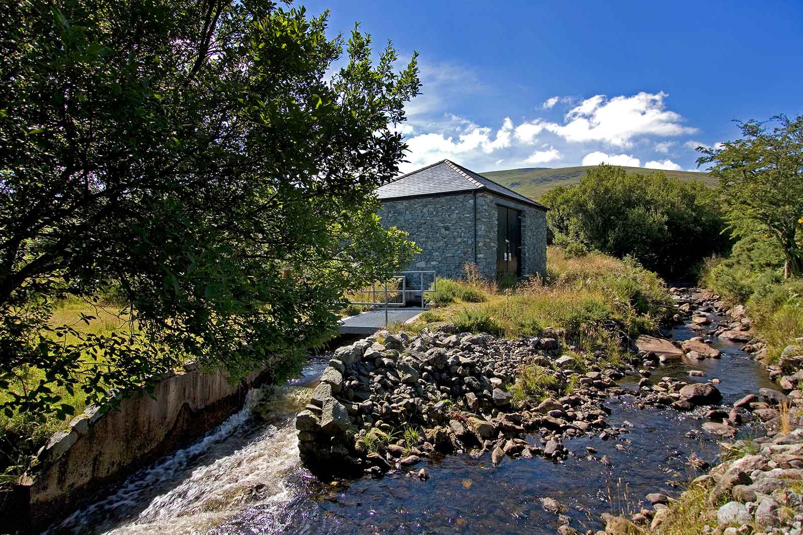 A stone building beside a flowing stream, surrounded by greenery, under a clear blue sky with fluffy clouds.