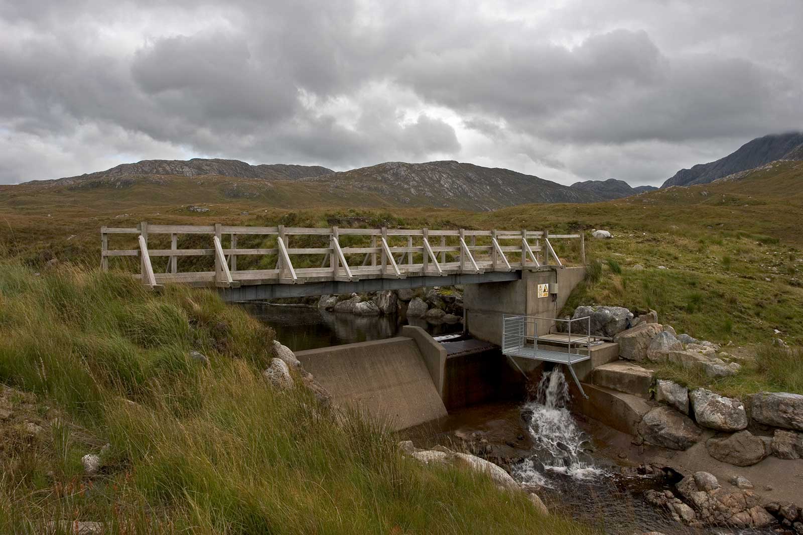 A wooden bridge crosses a stream, set against a backdrop of hills under a cloudy sky.
