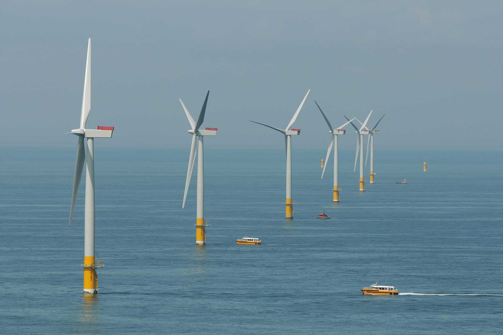 A row of offshore wind turbines with boats navigating in calm waters under a clear sky.