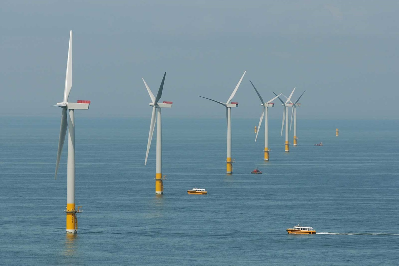 A row of offshore wind turbines with boats navigating in calm waters under a clear sky.