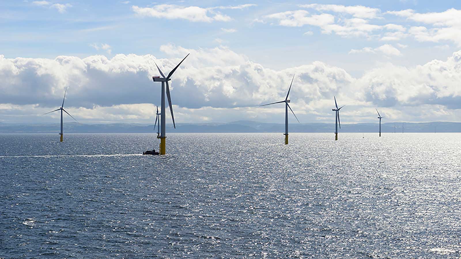 A scenic view of offshore wind turbines standing in the ocean, with a boat navigating the water under a cloudy sky.