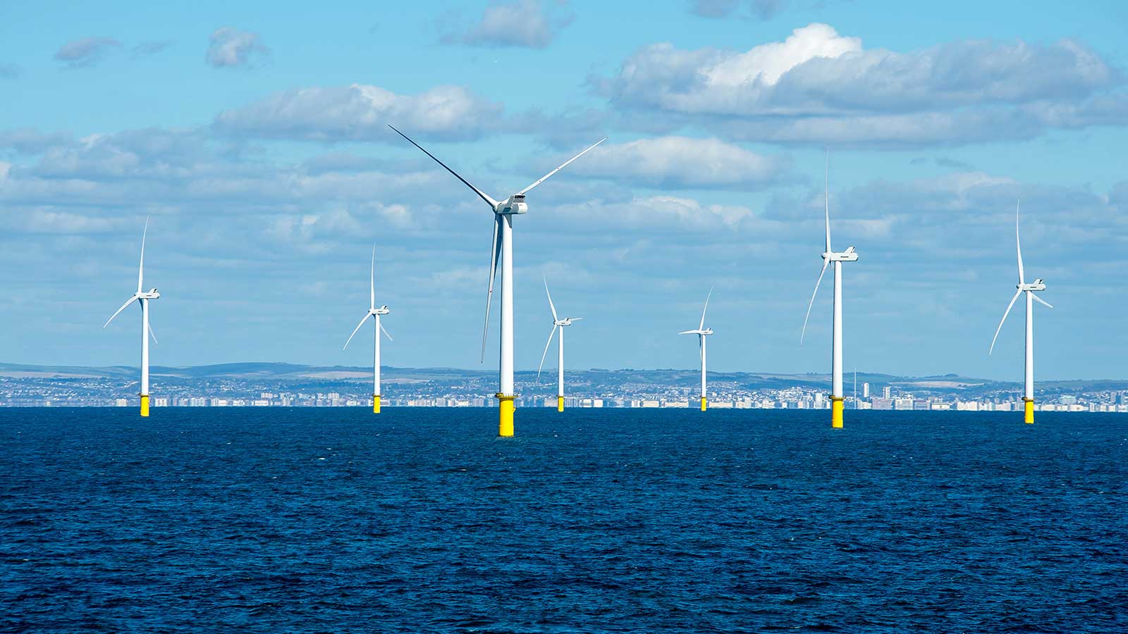 A series of wind turbines stand on yellow bases in the blue sea under a clear sky with clouds.