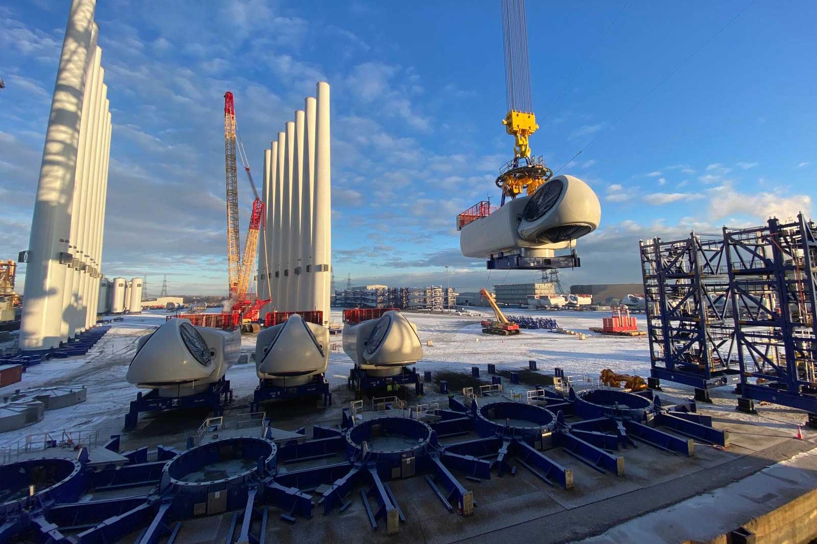 A crane lifts a wind turbine component on a snowy construction site with large concrete pillars and other machinery in the background.