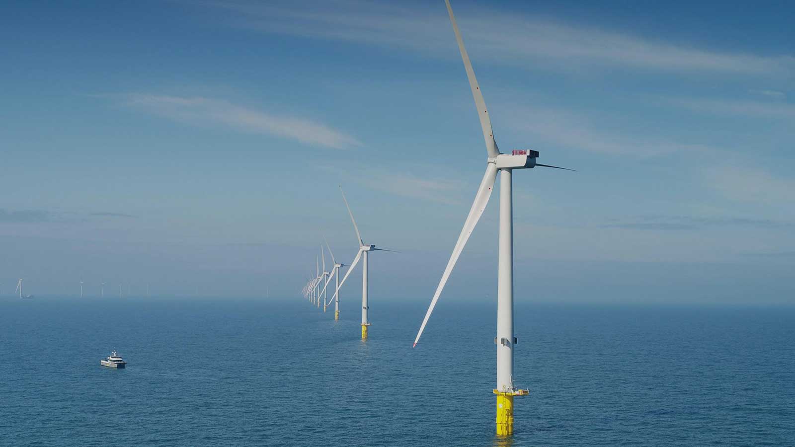 A row of offshore wind turbines stands tall in the ocean under a clear blue sky, with a small boat nearby.