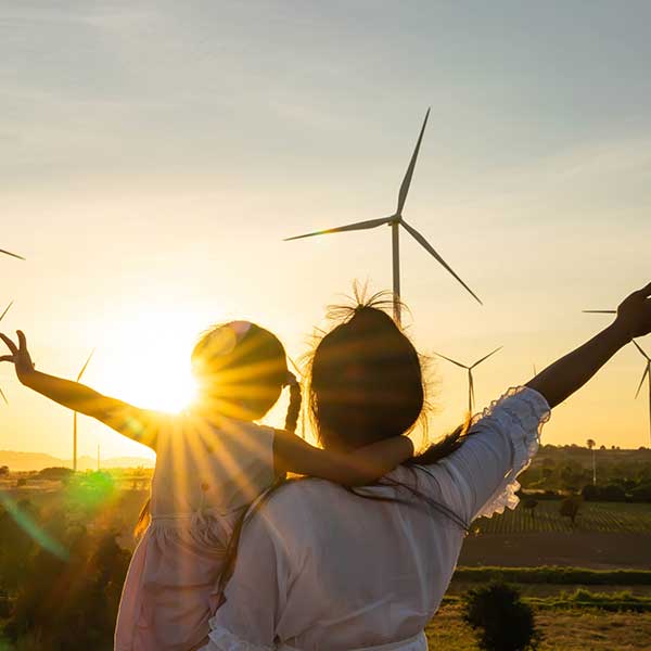 A silhouetted mother and daughter stand against a sunset, with wind turbines in the background, arms raised in joy.