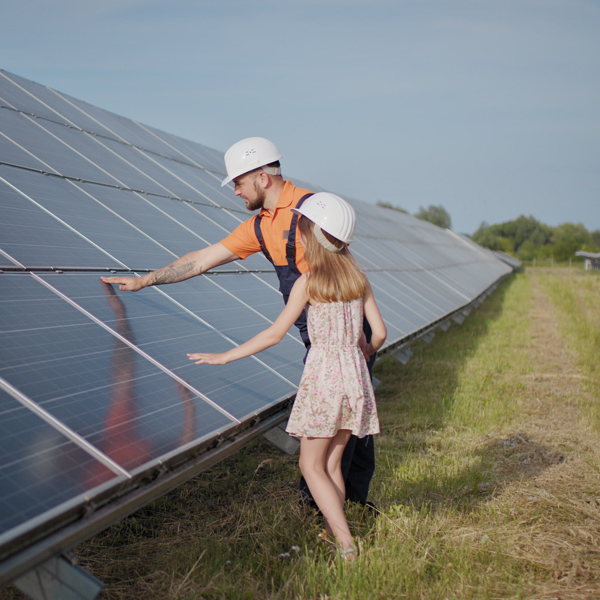 A person and a child examine solar panels in a field, both wearing safety helmets under a clear blue sky.