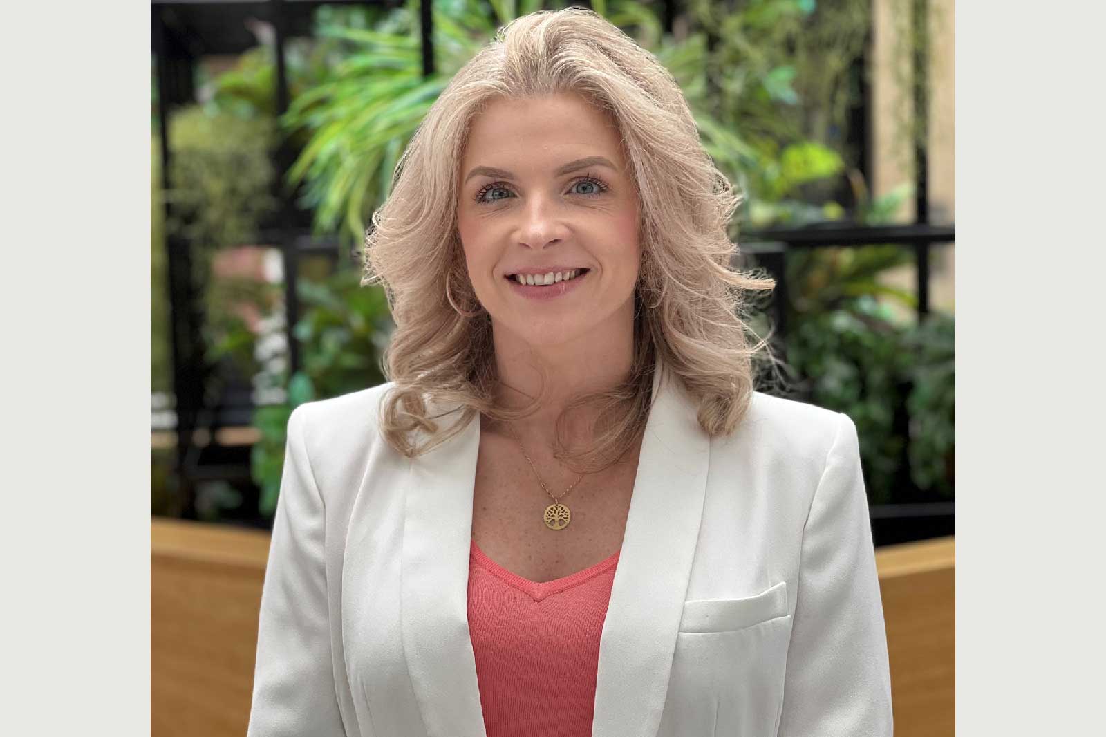 A woman with wavy blonde hair wears a white blazer over a pink top, standing in a leafy green environment.