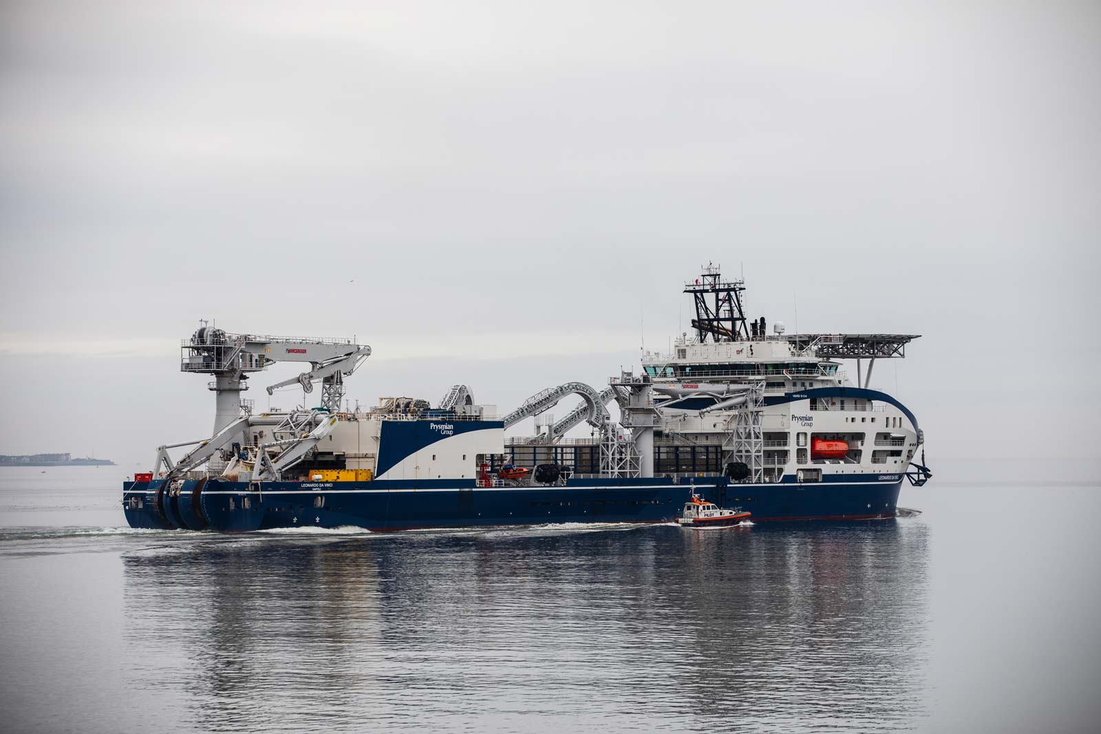 A large research vessel navigates calm waters, with a smaller boat nearby, under a cloudy sky.
