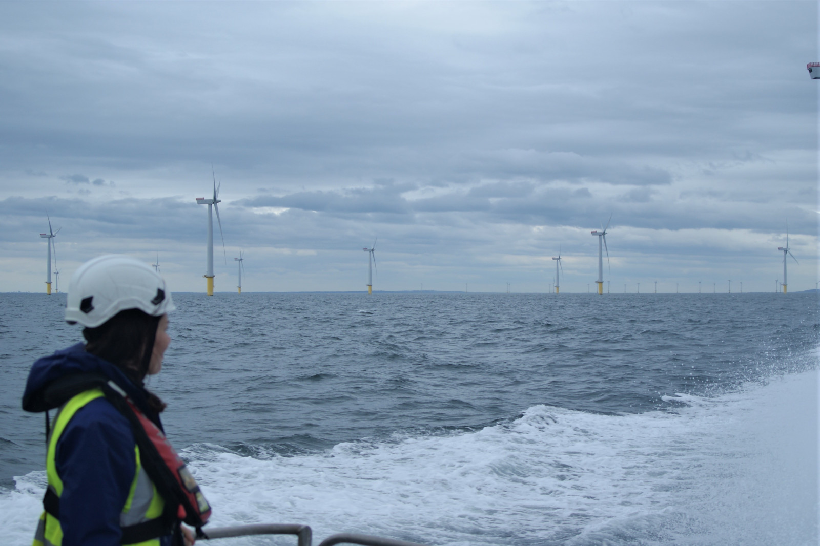 A person in safety gear observes offshore wind turbines in a cloudy sky while on a boat in choppy waters.