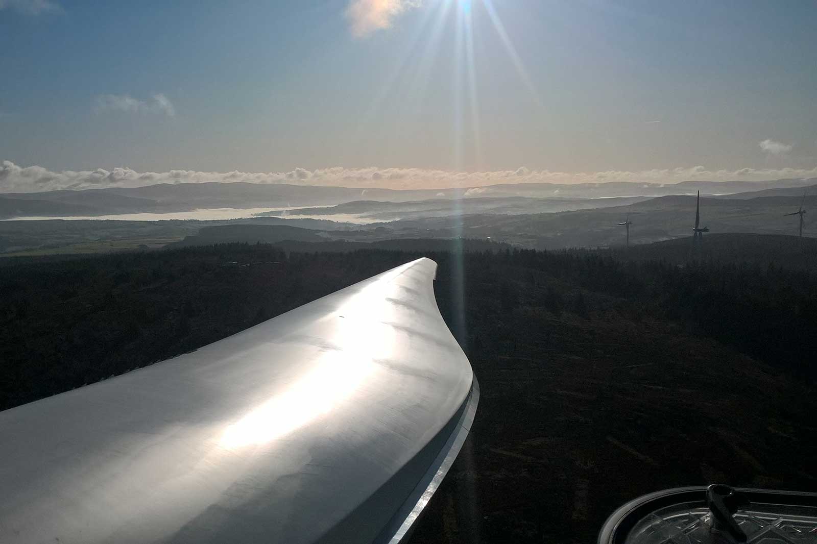 A close-up view of a wind turbine blade against a mountainous landscape under a bright sun, with distant hills and wind turbines visible.