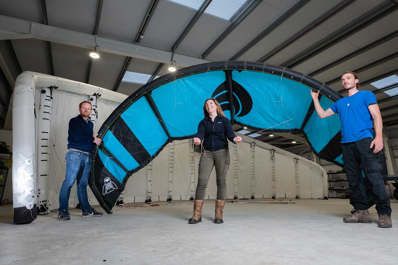 Three individuals in a warehouse hold a large blue and black kite, preparing for kiteboarding.