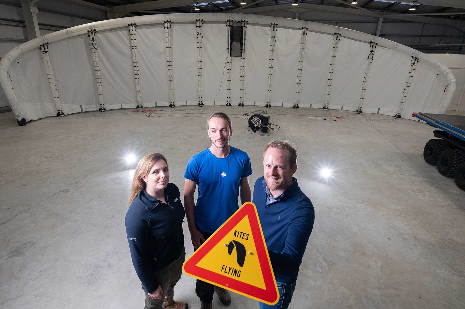 Three individuals hold a 'Kites Flying' warning sign in an indoor space with a large inflatable structure in the background.