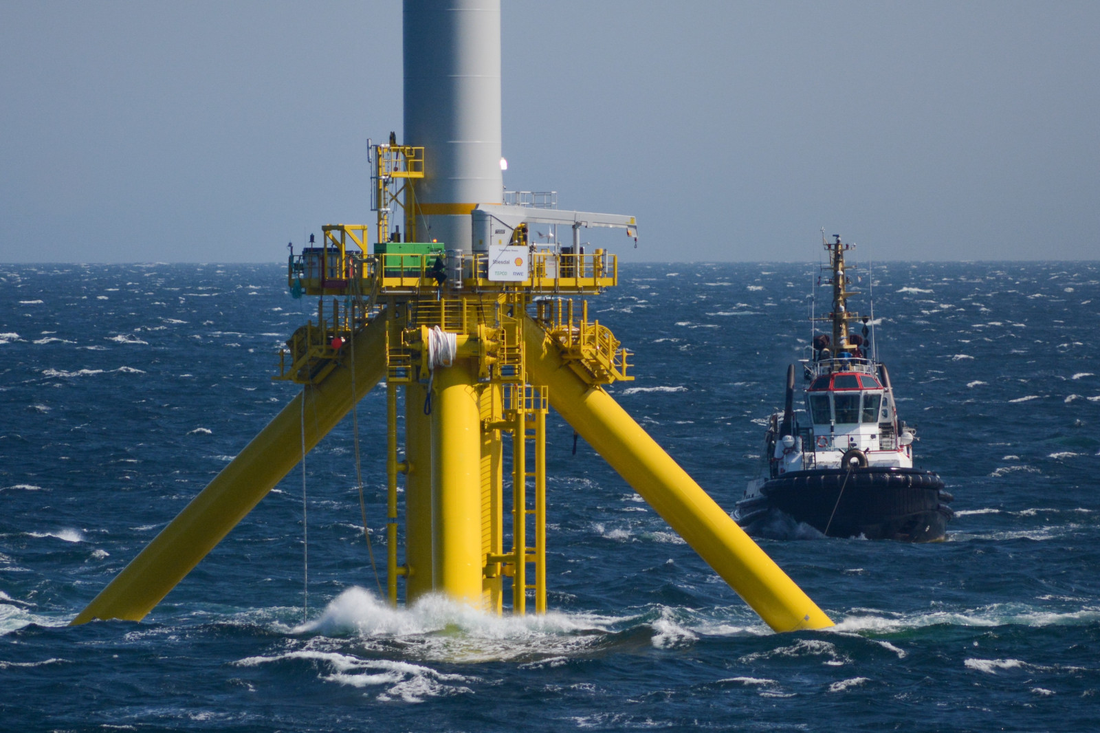 A yellow offshore wind turbine base stands in turbulent sea waters, with a tugboat approaching from the right.