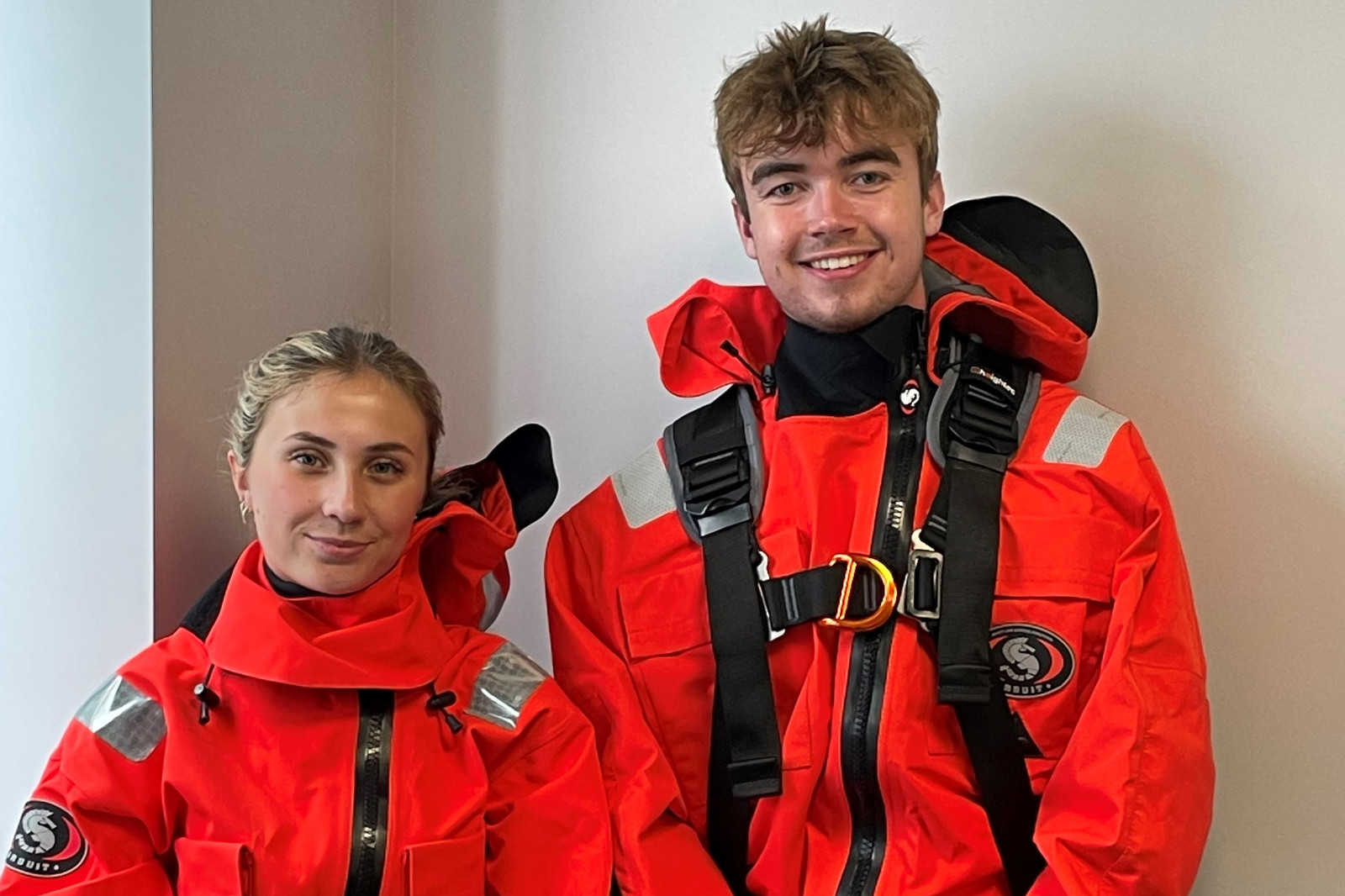 Two individuals in bright orange protective suits stand closely together, showcasing safety gear and equipment.