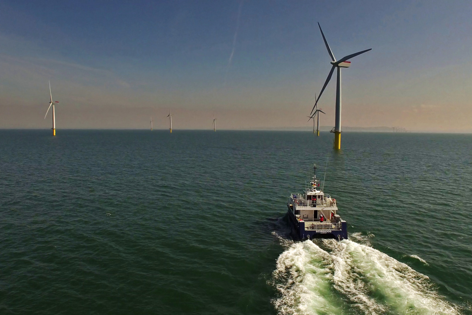 A boat navigates past several offshore wind turbines in a calm sea under a clear sky.