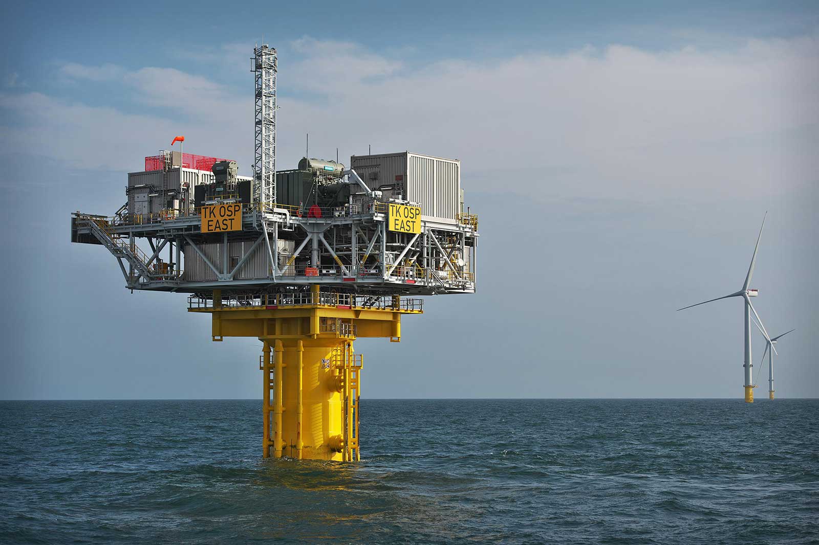 An offshore platform marked 'TK OSP EAST' with wind turbines in the background under a cloudy sky.