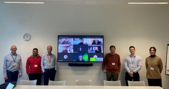A group of six people stands in a meeting room, with a large screen displaying remote participants. 