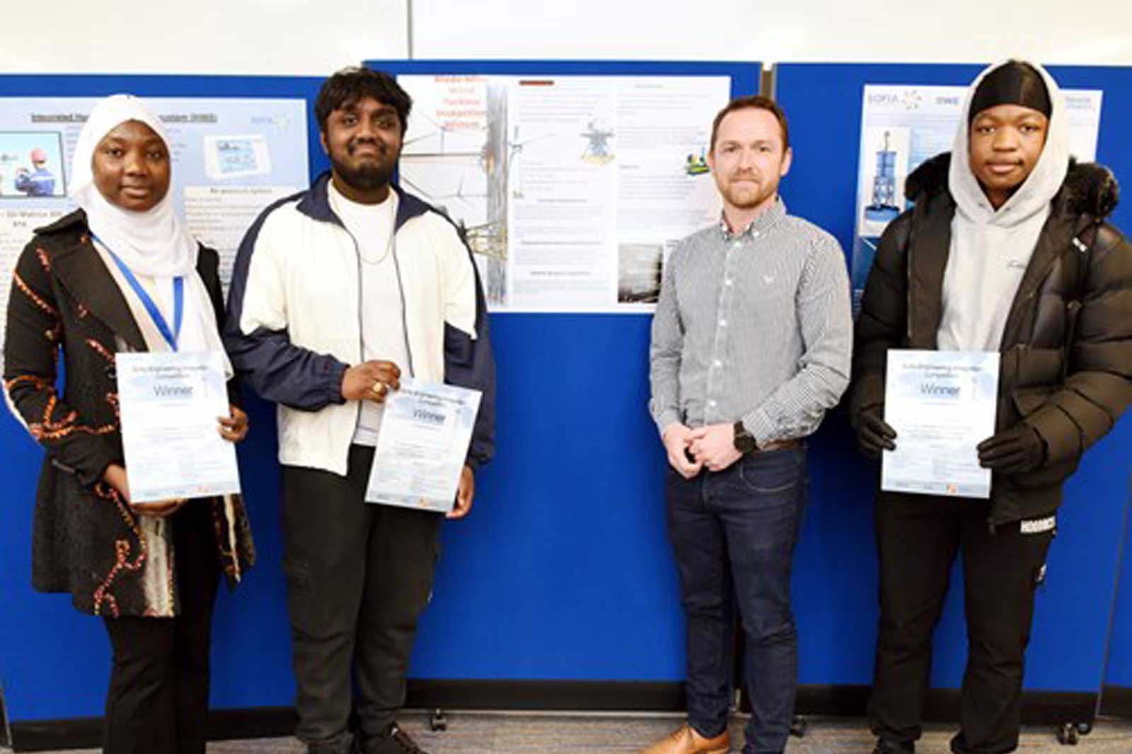 Four individuals stand together, holding certificates, in front of display boards with various informational posters.