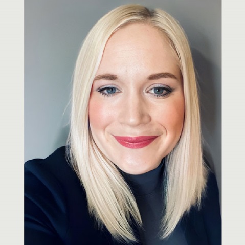 A person with long straight blonde hair wearing a black blazer against a grey background.