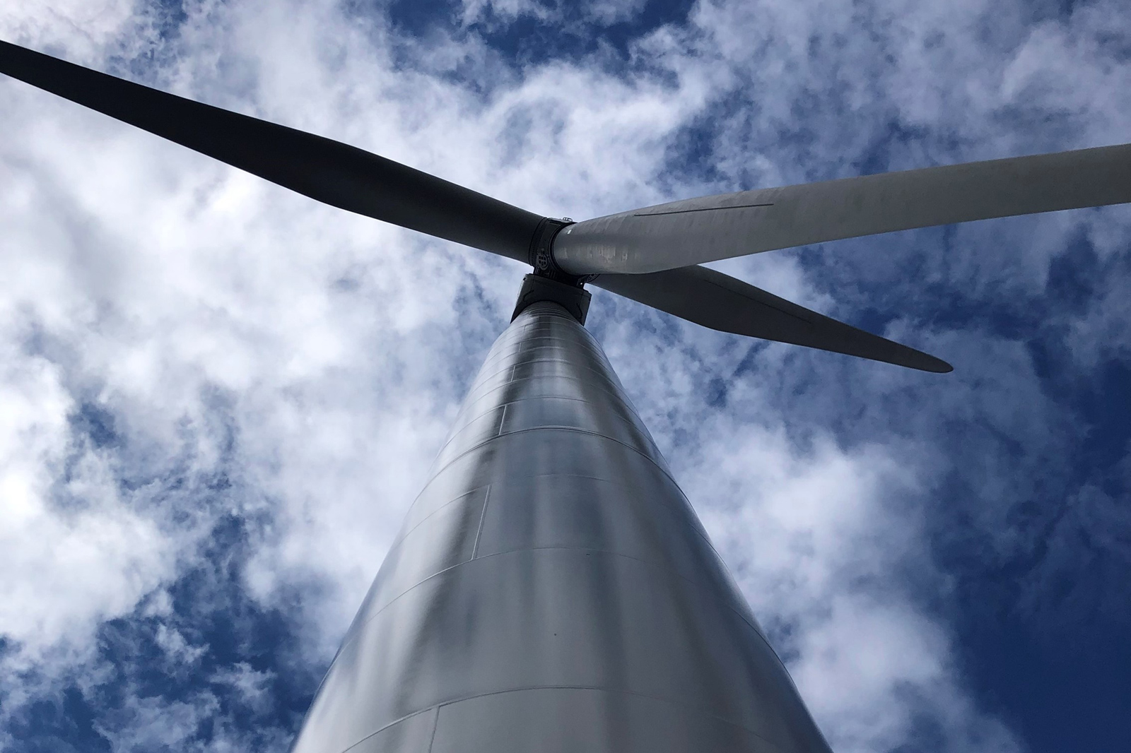 A view from below of a wind turbine. The tower is tall and white against a blue sky filled with white clouds.
