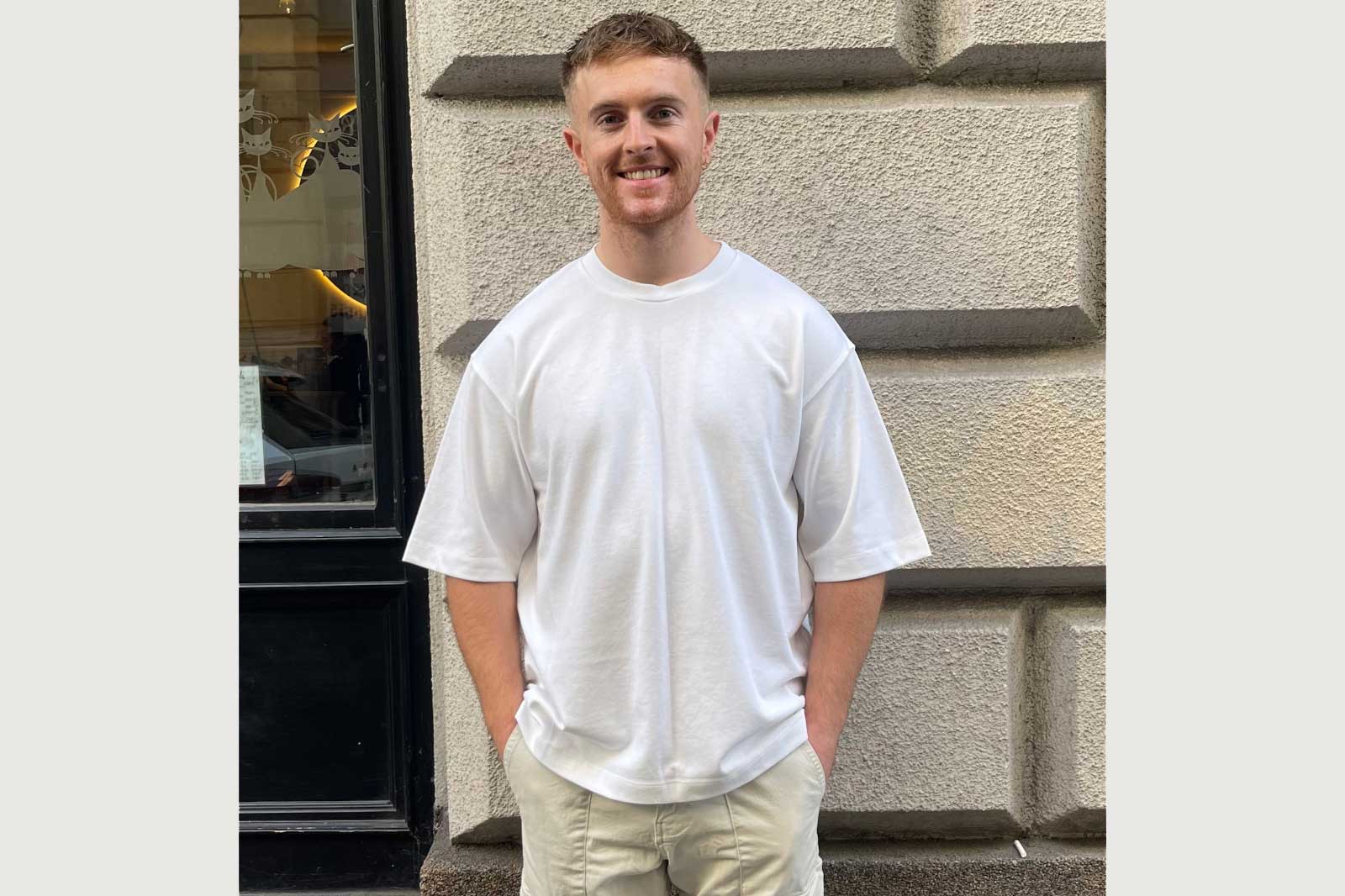 A person stands casually in a white t-shirt and shorts against a textured wall, hands in pockets.